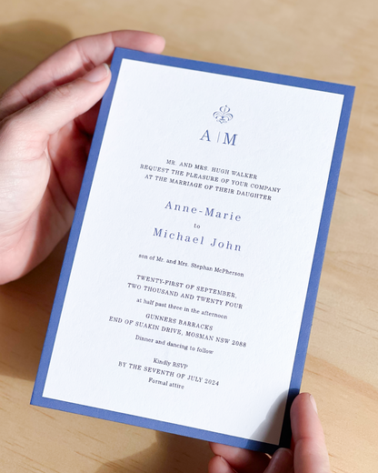 A close-up of a blue-bordered wedding invitation card with white paper and elegant blue text, printed with letterpress technology.